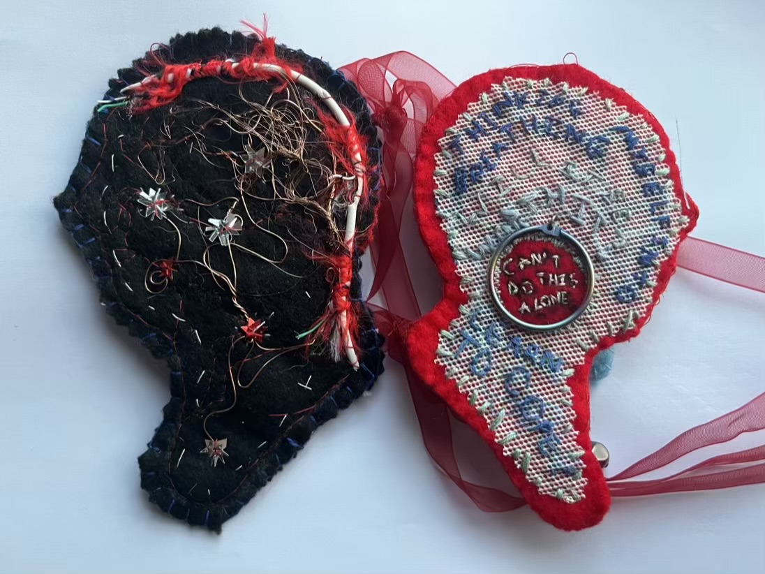 Two silhouettes of faces face away from each other. The one on the left is made of black felt with copper wires and shattered sequins stitched over it. Along the back of the head runs a red and white shredded charging cable. The right head is made of red felt with a layer of white plastic net over it. The words “thinking myself into breathing, will it be worth it, learn how to cope” stitched in blue thread. A iridescent hoop sits in the middle the words “can’t do this alone” stitched with white thread in the centre.