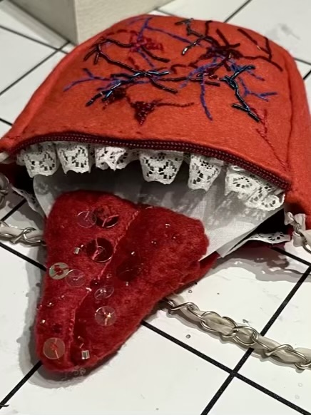 Embroidery of red and blue veins, some of them in metallic threads covers the top of the red felt bag. A red zip sits above pleated white lace teeth. Covered in shining beads and sequins a large red felt tongue sticks out from the bags opening.