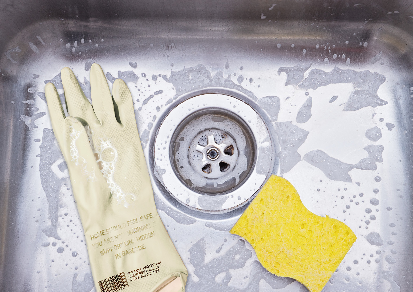 A close up photograph of a pair of Domestic Aid washing up gloves submerged in a soapy kitchen sink, with the hidden support message visible on the glove surface.